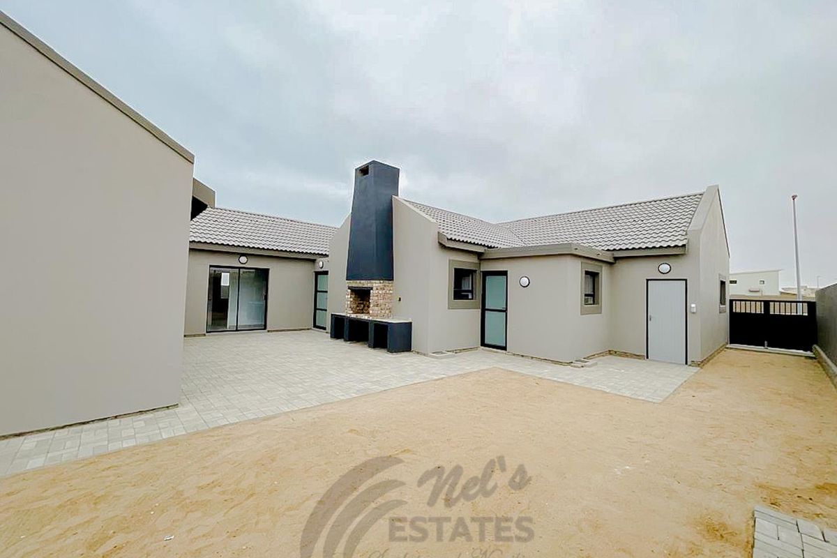 A modern rear exterior view featuring a paved courtyard, a built-in outdoor fireplace, and neutral-toned exterior walls with tiled roofing.