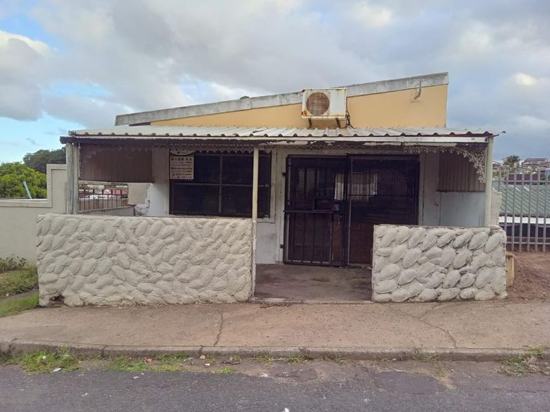 Front elevation of a single-story property featuring a stone-clad low wall, metal security gate, and corrugated roof structure in need of maintenance.