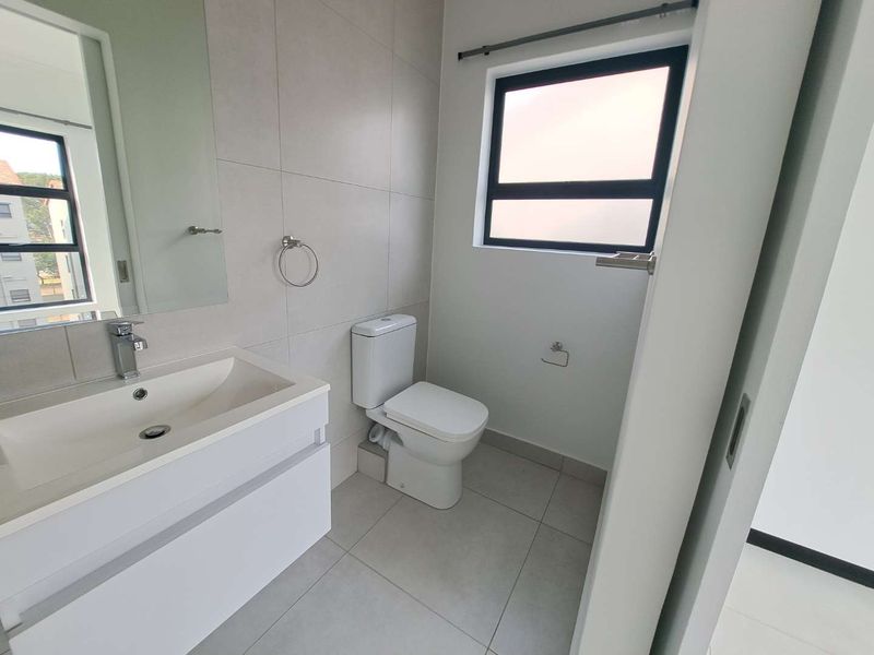 A modern, clean bathroom featuring a sleek vanity cabinet, ceramic basin, toilet, and neutral wall and floor tiling with a frosted window.