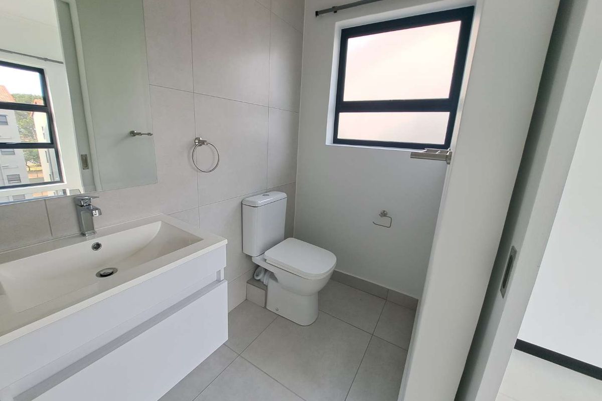 A modern, clean bathroom featuring a sleek vanity cabinet, ceramic basin, toilet, and neutral wall and floor tiling with a frosted window.