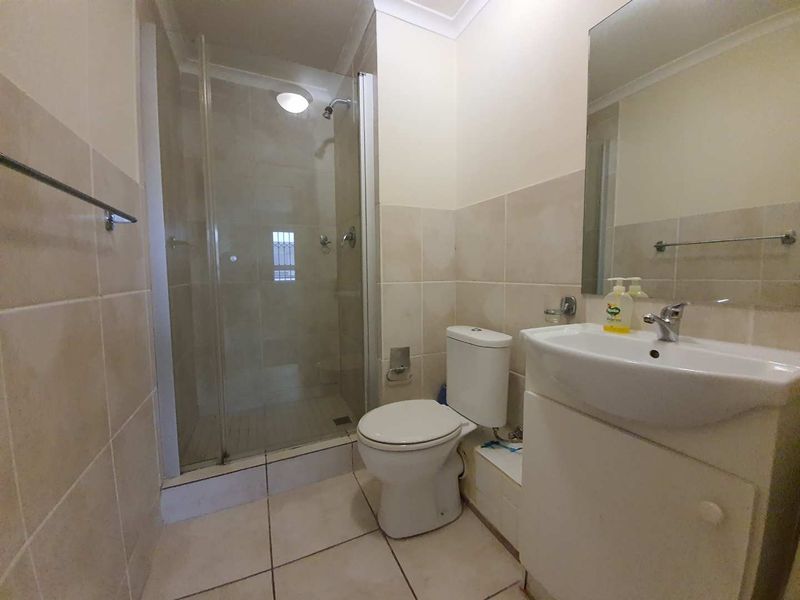 A functional bathroom featuring a glass-enclosed shower, a white vanity basin with a wall-mounted mirror, and neutral floor-to-ceiling wall tiles.