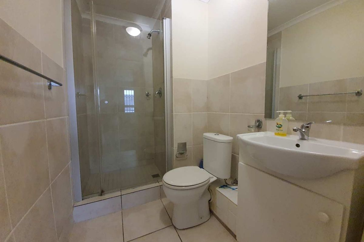 A functional bathroom featuring a glass-enclosed shower, a white vanity basin with a wall-mounted mirror, and neutral floor-to-ceiling wall tiles.