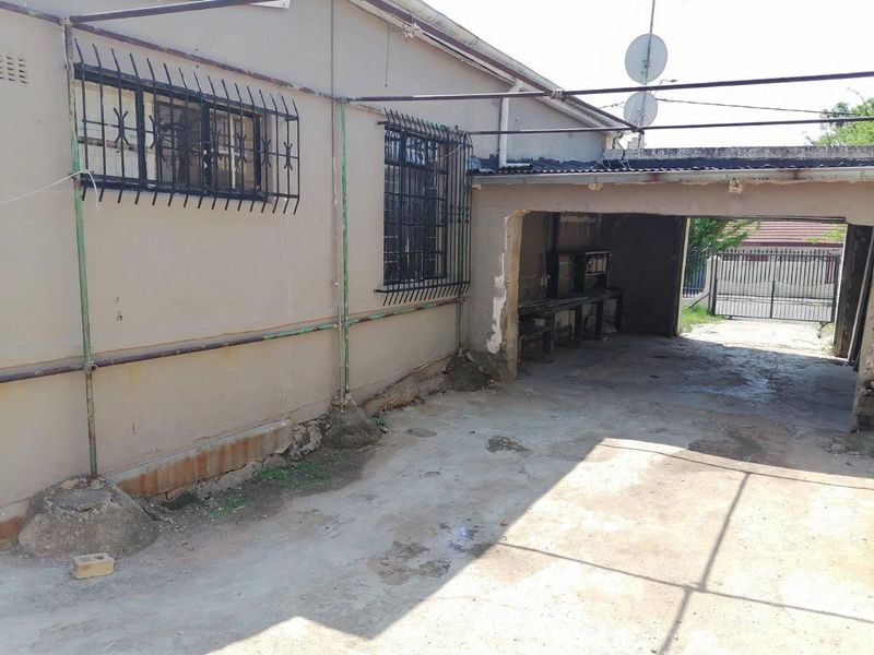 Rear exterior view showing a concrete driveway, carport structure, and security bars on windows, requiring maintenance and cosmetic repairs.