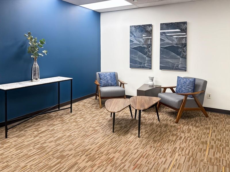 A professional office waiting area featuring a blue accent wall, recessed ceiling lighting, and neutral patterned carpeting.