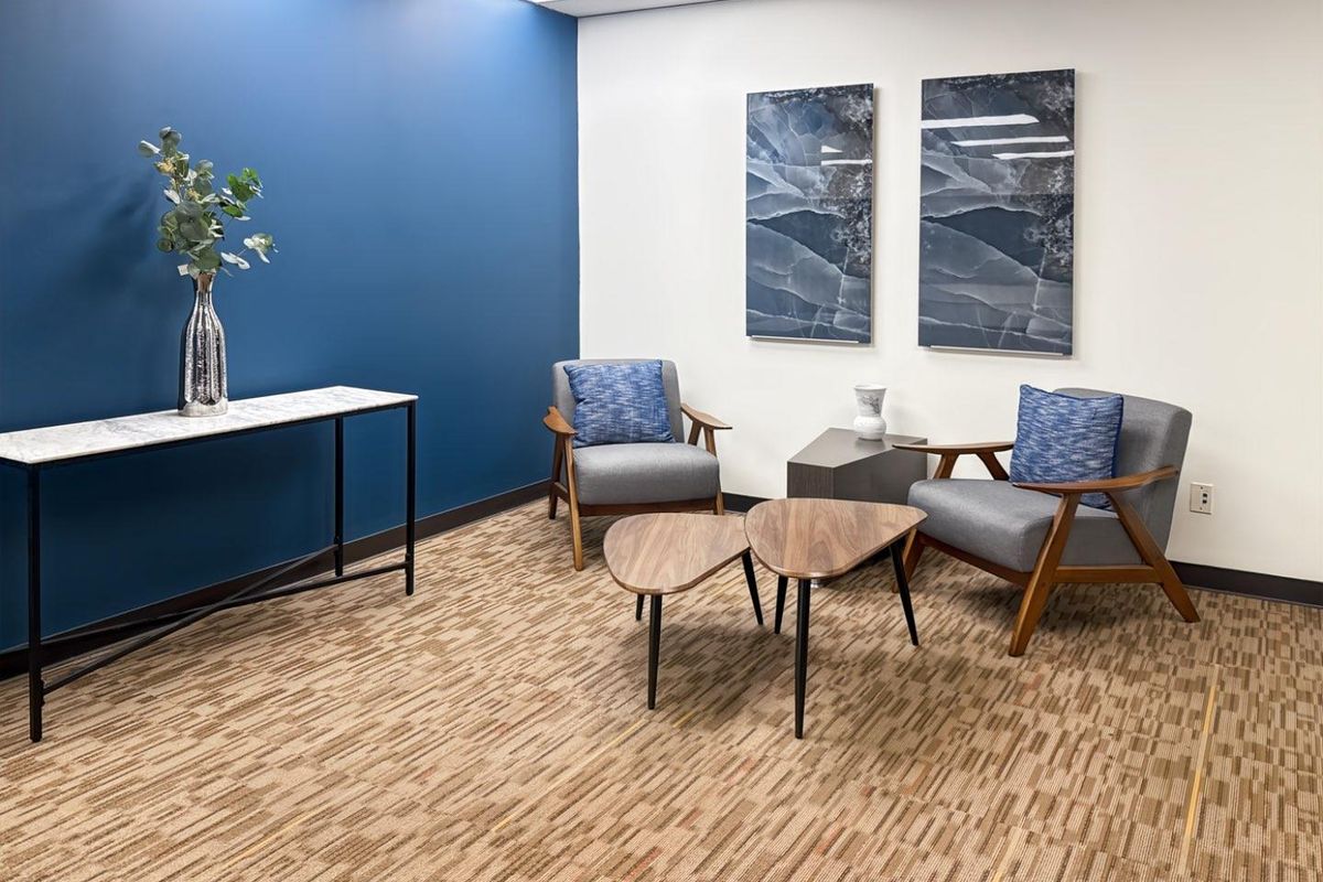 A professional office waiting area featuring a blue accent wall, recessed ceiling lighting, and neutral patterned carpeting.