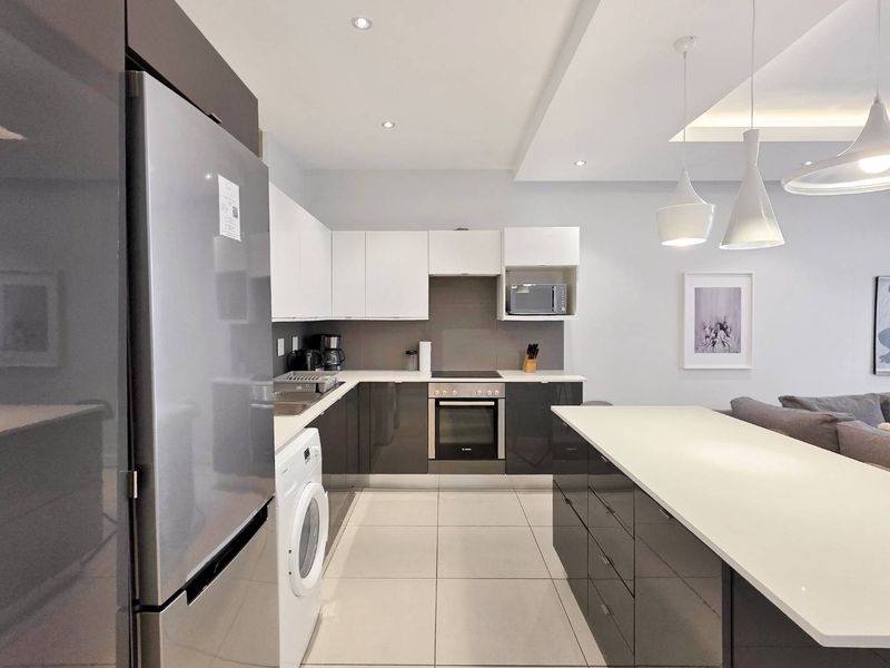 Modern open-plan kitchen featuring high-gloss cabinetry, integrated appliances, a central island, and sleek tiled flooring, seamlessly connecting to the living area.