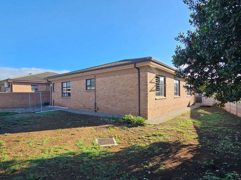 Rear view of the brick house featuring a tiled roof, louvered windows, and a paved walkway surrounding the structure with a garden area.