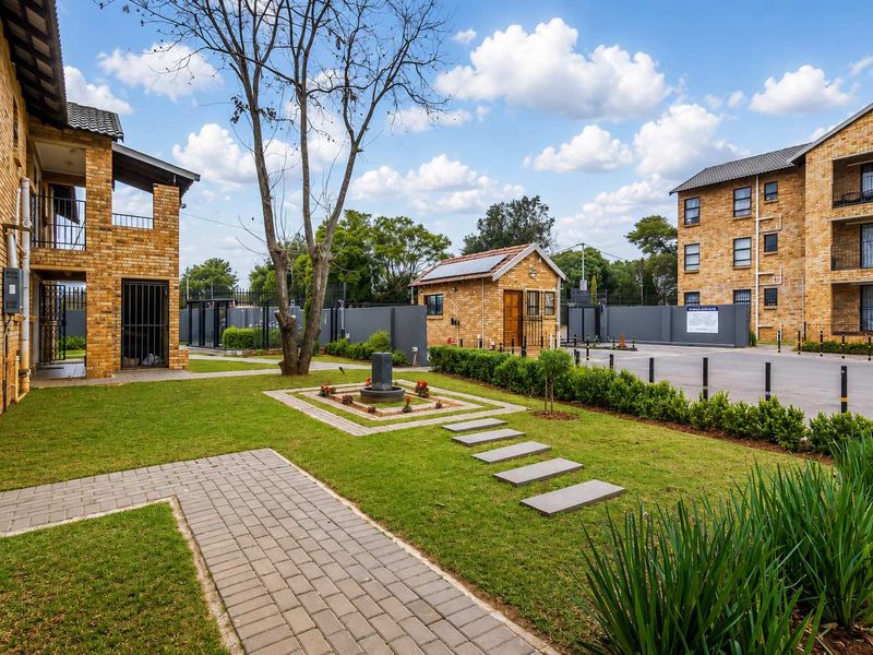 A well-maintained apartment complex exterior featuring brick facades, landscaped gardens, paved walkways, and a secure gated entrance with a guard house.