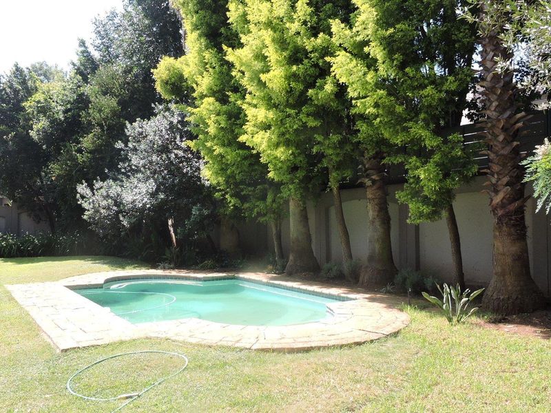 A private backyard featuring a swimming pool with stone coping, surrounded by a lush garden and established trees against a boundary wall.
