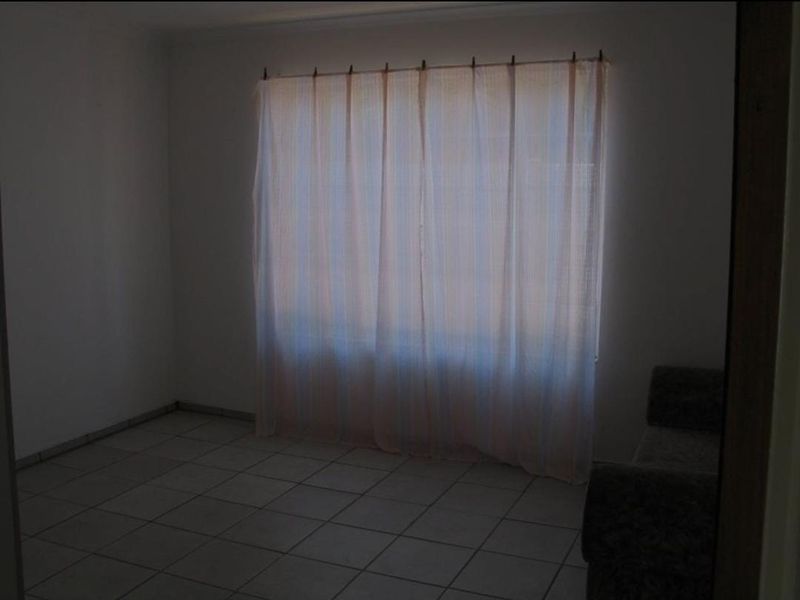 A sparsely furnished living area featuring neutral tiled flooring and a large window with curtains, offering a blank canvas for interior design.