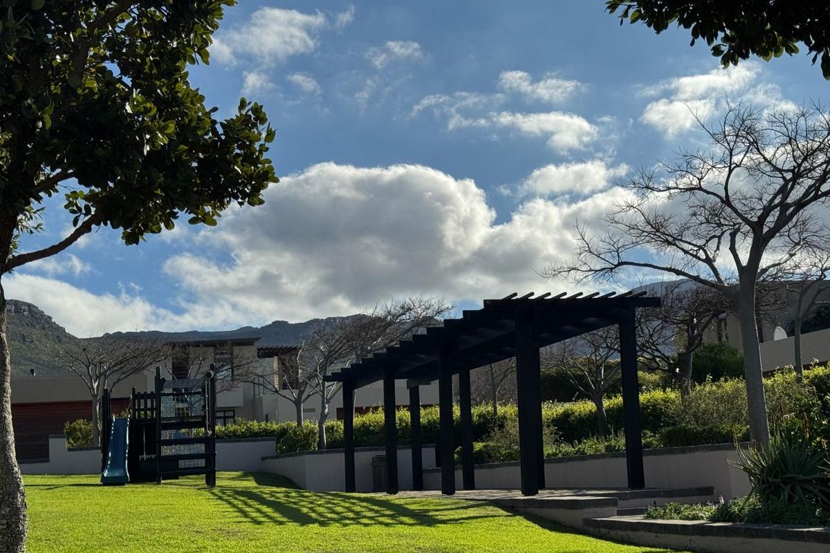 A well-maintained communal garden area featuring a modern pergola, manicured lawn, and retaining walls with mountain views in the background.