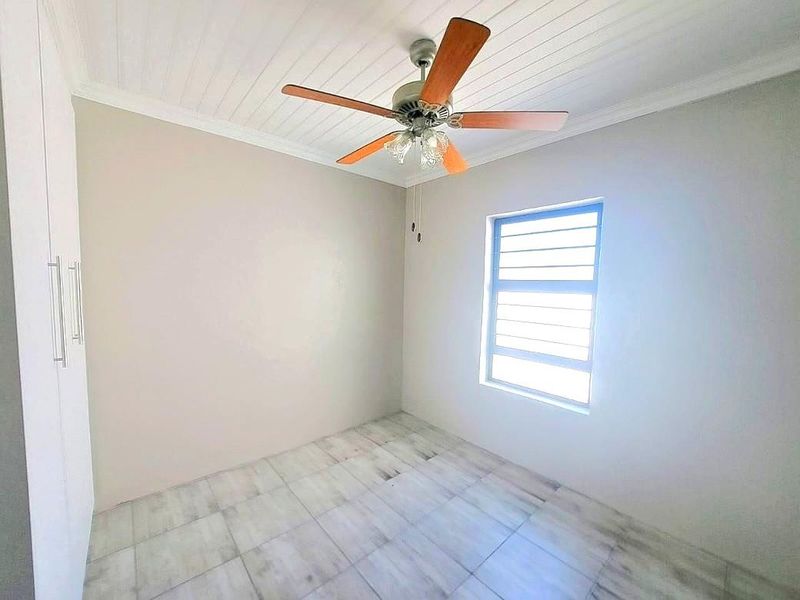 A bright, empty bedroom featuring tiled flooring, built-in cupboards, a ceiling fan, and a louvered window.