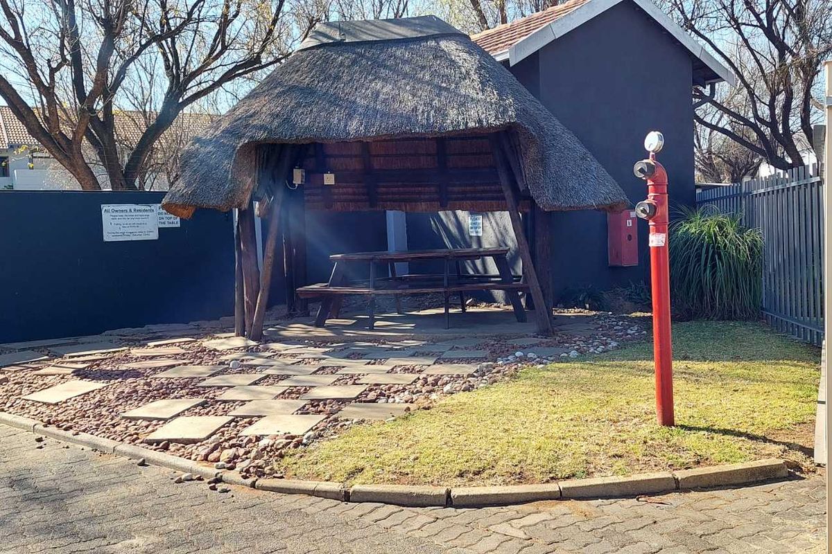 A communal outdoor area featuring a traditional thatch lapa, paved ground, and a fire hydrant within a secure apartment complex setting.