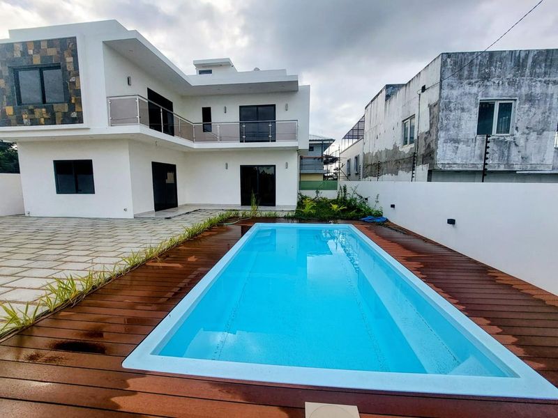 Modern two-story house exterior featuring a rectangular swimming pool with timber decking, glass balcony railings, and paved patio area.