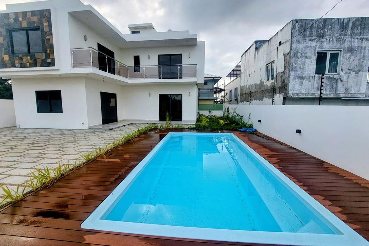 Modern two-story house exterior featuring a rectangular swimming pool with timber decking, glass balcony railings, and paved patio area.