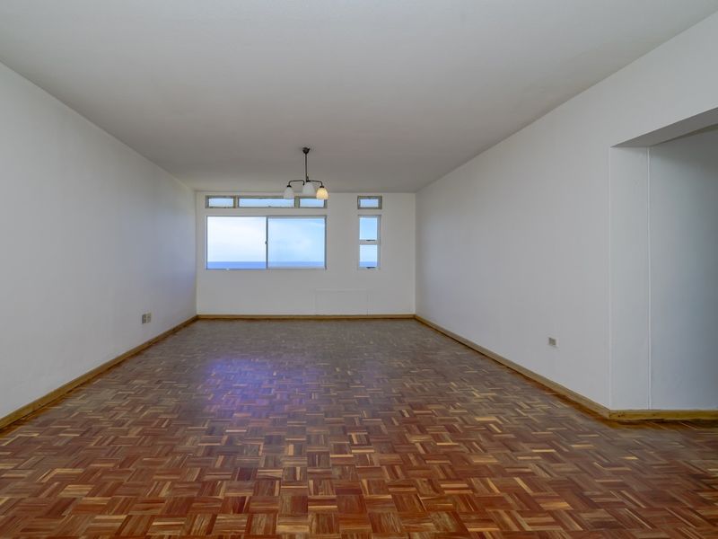 Spacious, empty living area featuring classic parquet flooring, neutral white walls, and large windows offering natural light and ocean views.