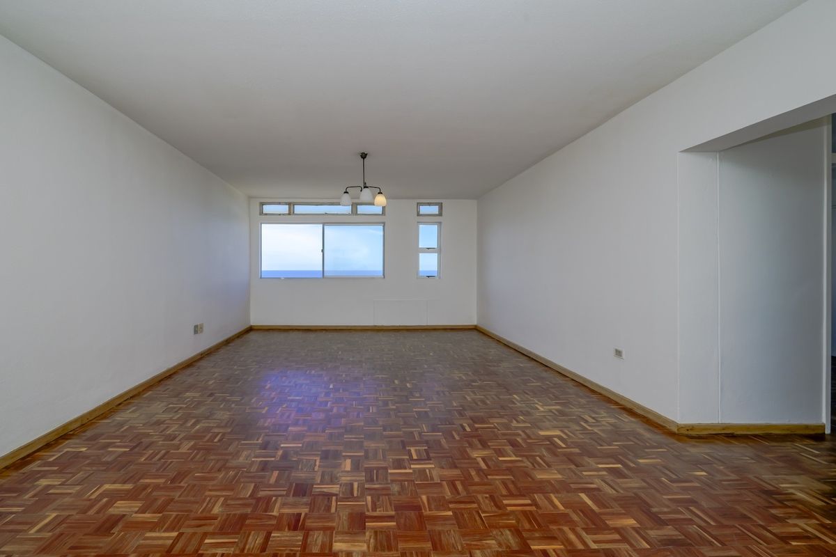 Spacious, empty living area featuring classic parquet flooring, neutral white walls, and large windows offering natural light and ocean views.