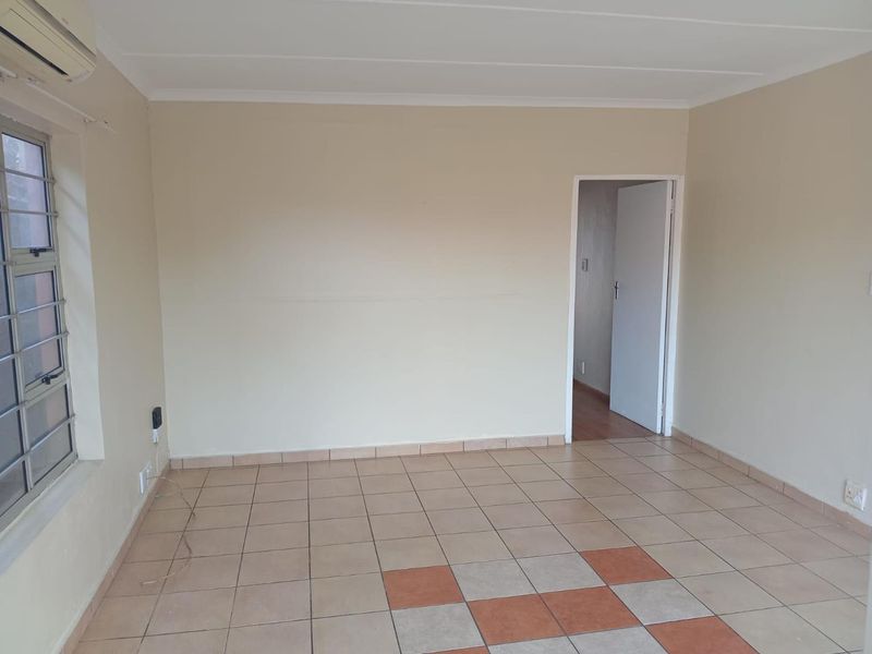 An empty living room featuring tiled flooring, an air conditioning unit, and security bars on the window, leading to an adjacent room.