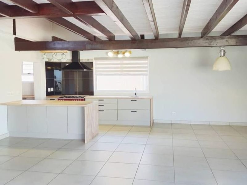 Spacious open-plan living area featuring exposed timber ceiling beams, modern kitchen cabinetry, a central island, and neutral tiled flooring throughout.