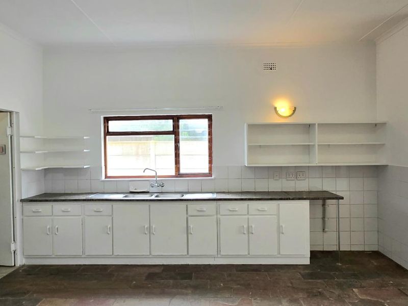 A spacious, empty kitchen featuring white built-in cabinetry, a stainless steel sink, tiled backsplash, and an adjacent built-in fireplace area.