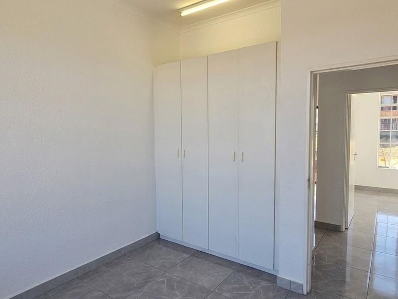 A clean, empty bedroom featuring built-in cupboards, neutral tiled flooring, and a doorway leading to an adjacent room.