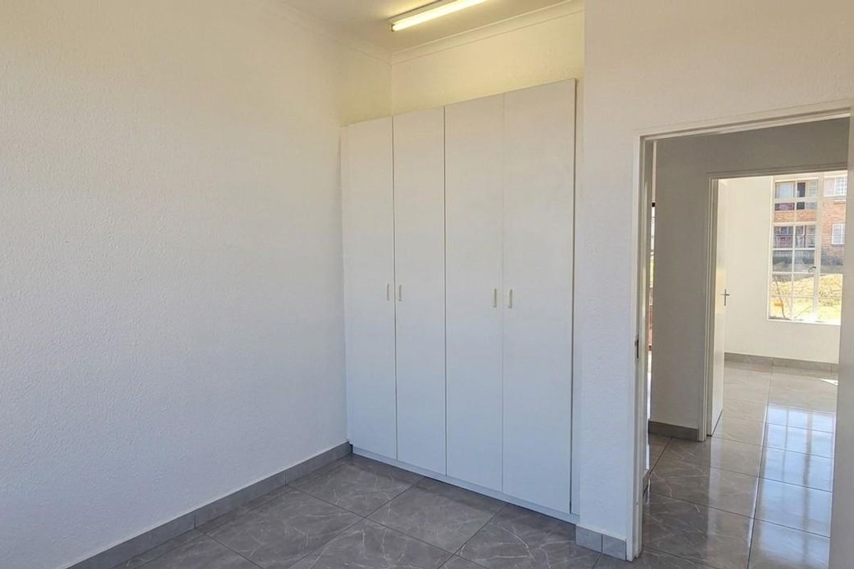 A clean, empty bedroom featuring built-in cupboards, neutral tiled flooring, and a doorway leading to an adjacent room.