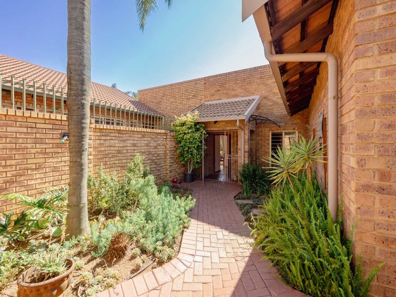 A well-maintained front entrance featuring a paved walkway, face brick exterior, and secure gated access, surrounded by established garden beds.