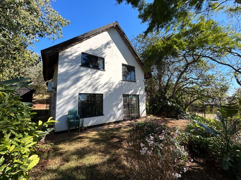 Rear view of a two-story cottage featuring white exterior walls, dark-framed windows with security bars, and a thatched roof set within a garden.