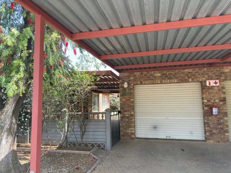 A covered carport area featuring a corrugated metal roof, brick facade, and a secure roller shutter garage door with paved driveway access.
