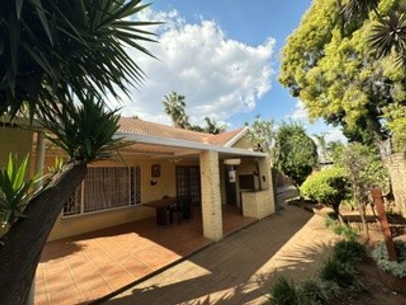 A covered outdoor patio area featuring tiled flooring, brick support pillars, and a built-in braai, overlooking the garden.