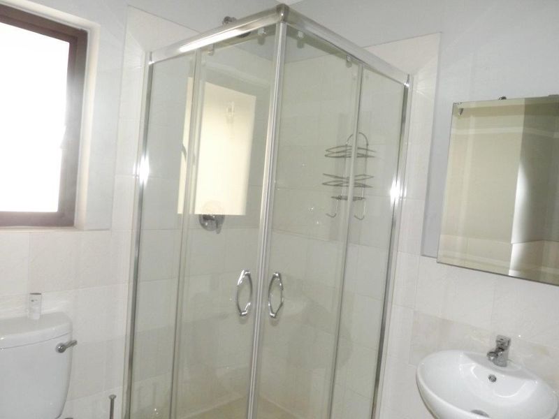 A clean, functional bathroom featuring a glass shower enclosure, pedestal basin, wall mirror, and neutral wall tiling.