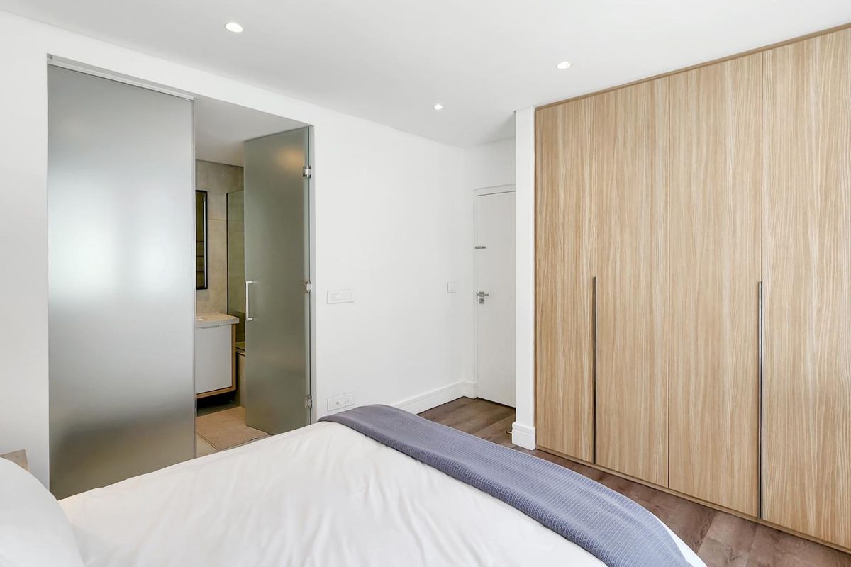Modern bedroom featuring built-in light wood cabinetry, recessed ceiling lighting, and an en-suite bathroom accessed through a contemporary frosted glass door.