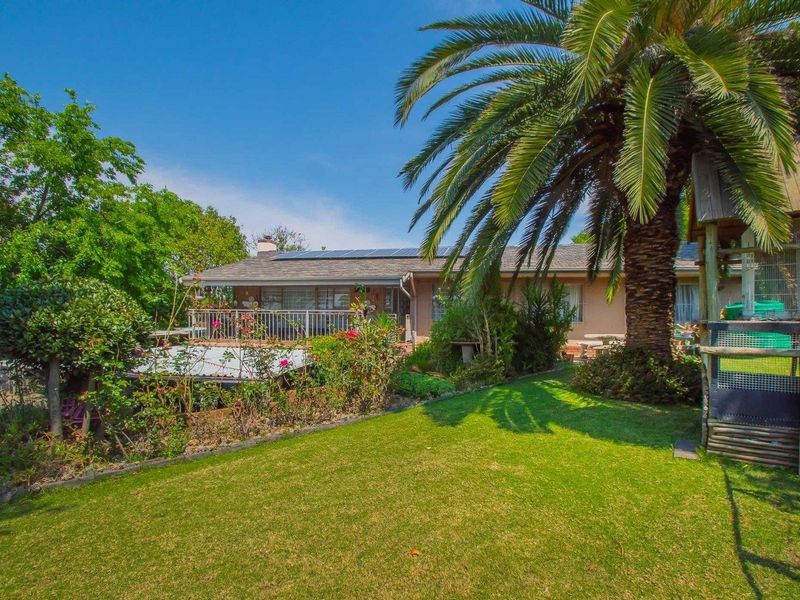 Rear view of the property featuring a landscaped garden, mature palm tree, and a raised deck with railing, complemented by roof-mounted solar panels.