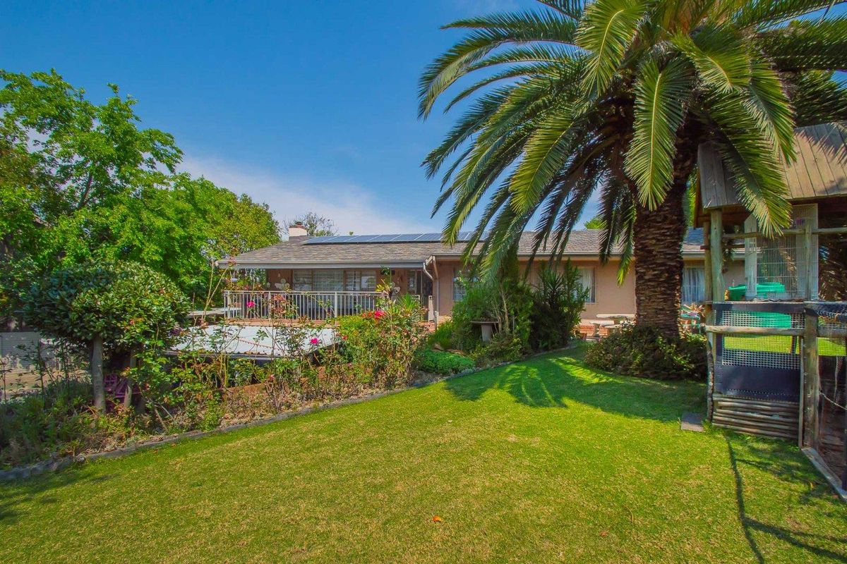 Rear view of the property featuring a landscaped garden, mature palm tree, and a raised deck with railing, complemented by roof-mounted solar panels.