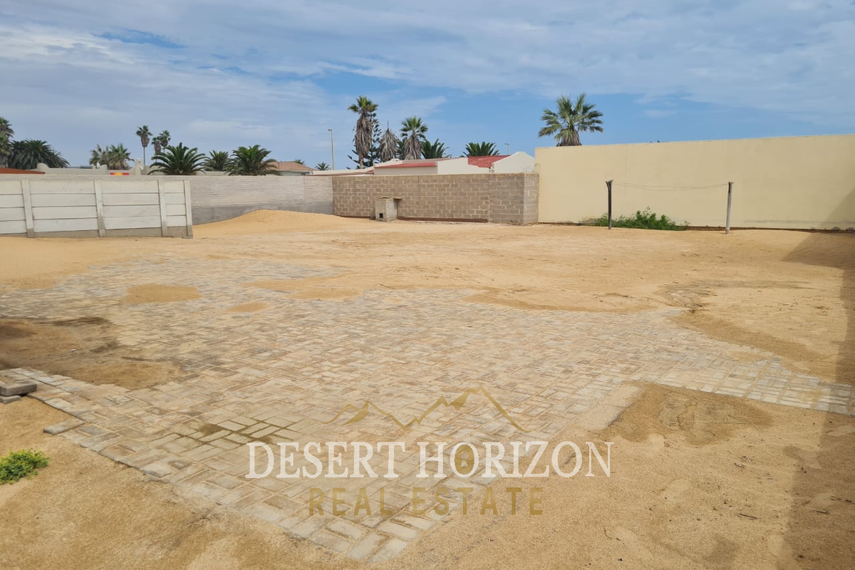A spacious, undeveloped rear yard featuring partial paving, sandy ground, and perimeter boundary walls, offering significant potential for landscaping or outdoor development.