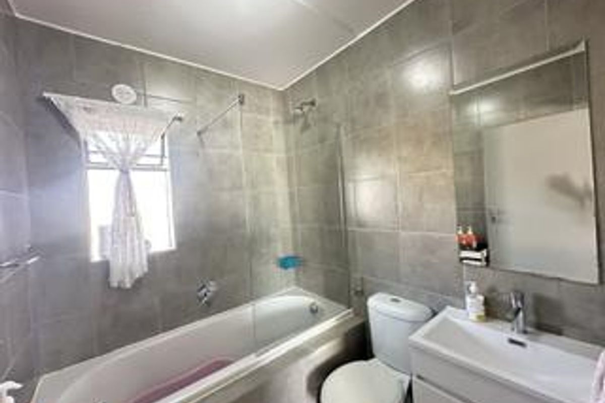 A modern bathroom featuring grey wall tiles, a bathtub with a glass shower screen, a wall-mounted vanity, and a toilet.
