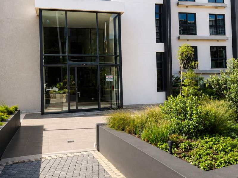 Modern retail building entrance featuring a glass facade, paved walkway, and well-maintained raised concrete planter boxes.