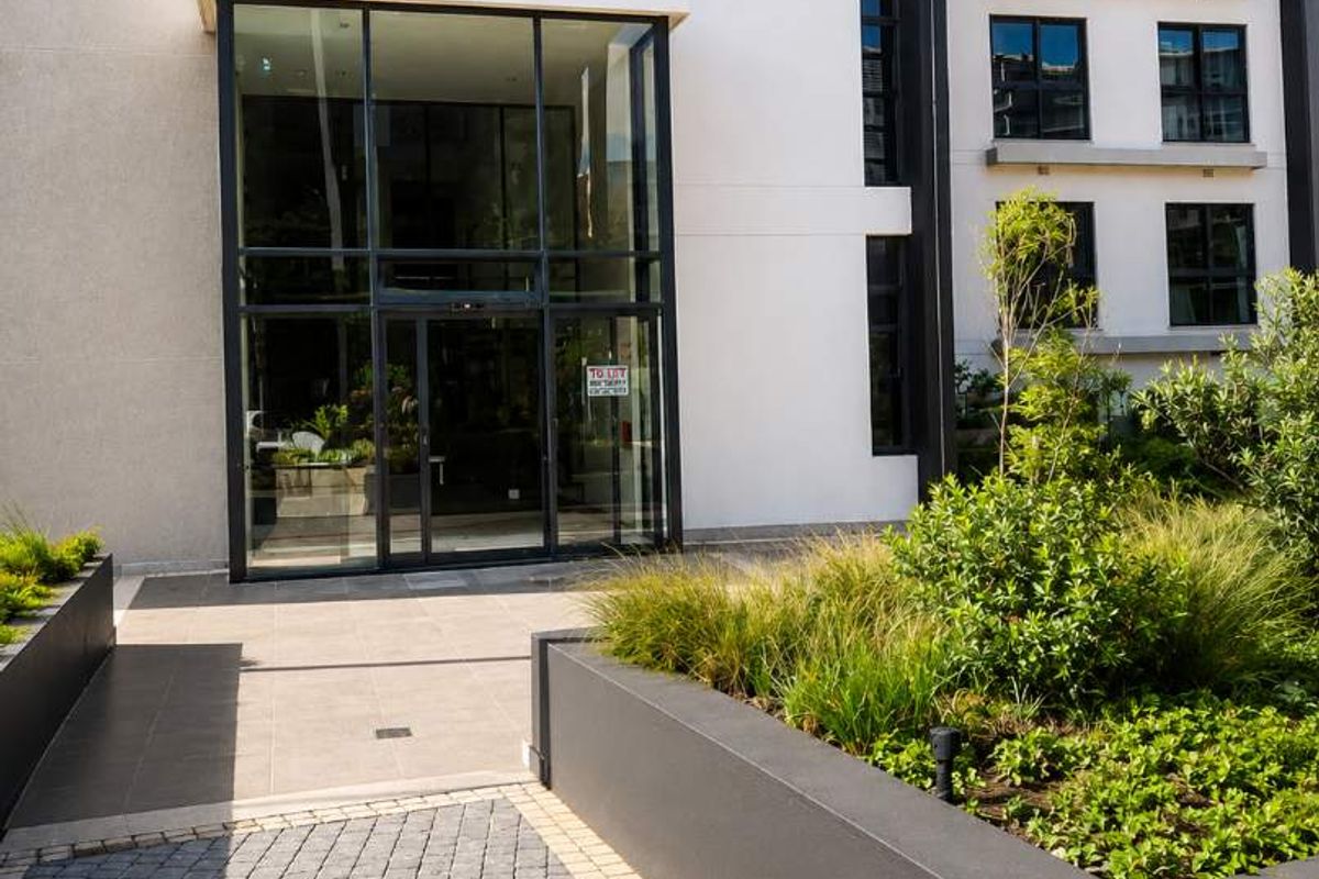 Modern retail building entrance featuring a glass facade, paved walkway, and well-maintained raised concrete planter boxes.