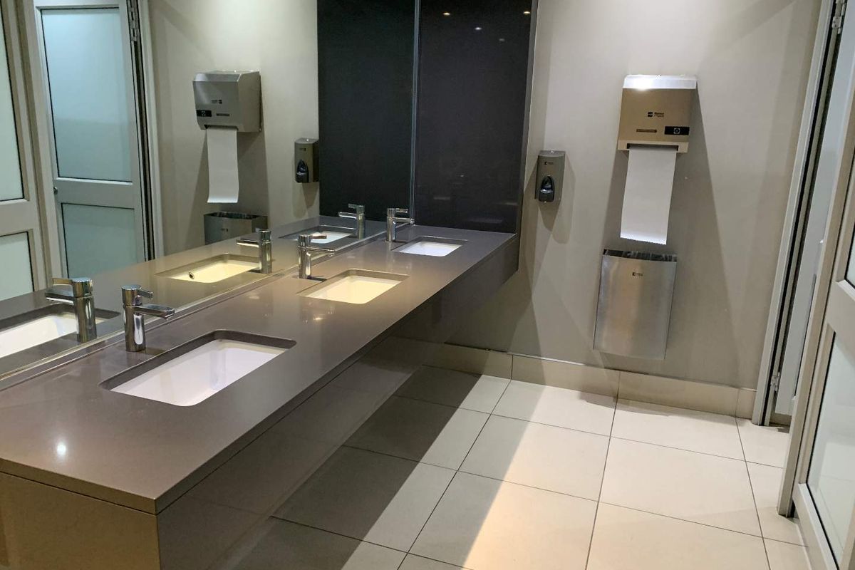 Modern office restroom featuring a floating vanity with dual integrated sinks, large wall-mounted mirror, and stainless steel commercial fixtures.