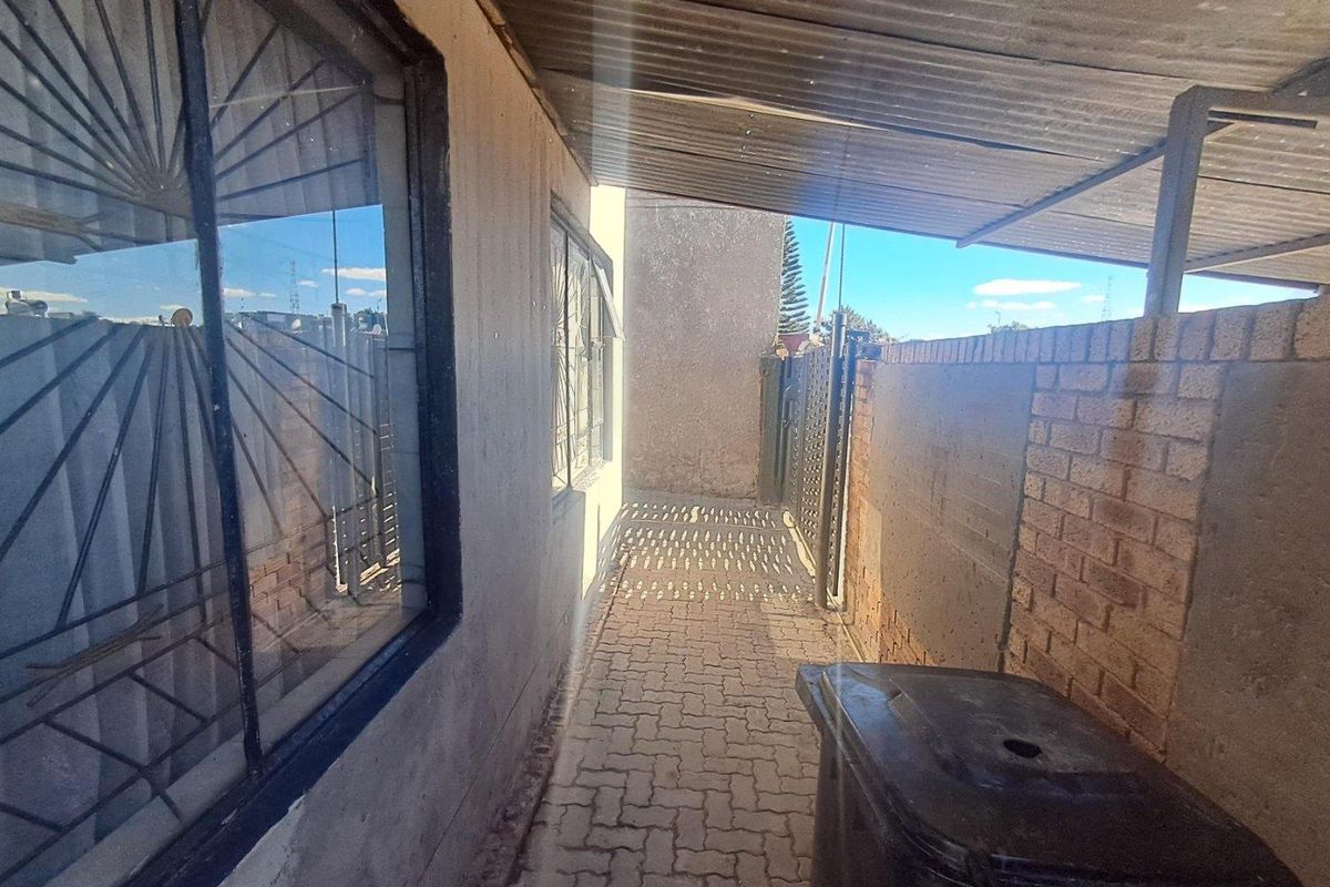 A narrow exterior side passage featuring paved flooring, a corrugated metal roof covering, and security bars on the windows.