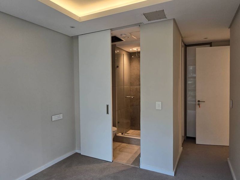 A clean, modern bedroom featuring neutral walls, carpet flooring, a recessed ceiling, and a sliding door leading to an en-suite bathroom with a glass shower.