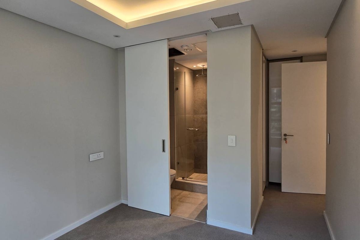 A clean, modern bedroom featuring neutral walls, carpet flooring, a recessed ceiling, and a sliding door leading to an en-suite bathroom with a glass shower.