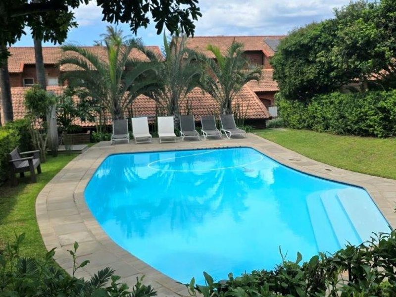 A well-maintained outdoor swimming pool area featuring a paved surround, integrated steps, and established garden landscaping within a residential complex.