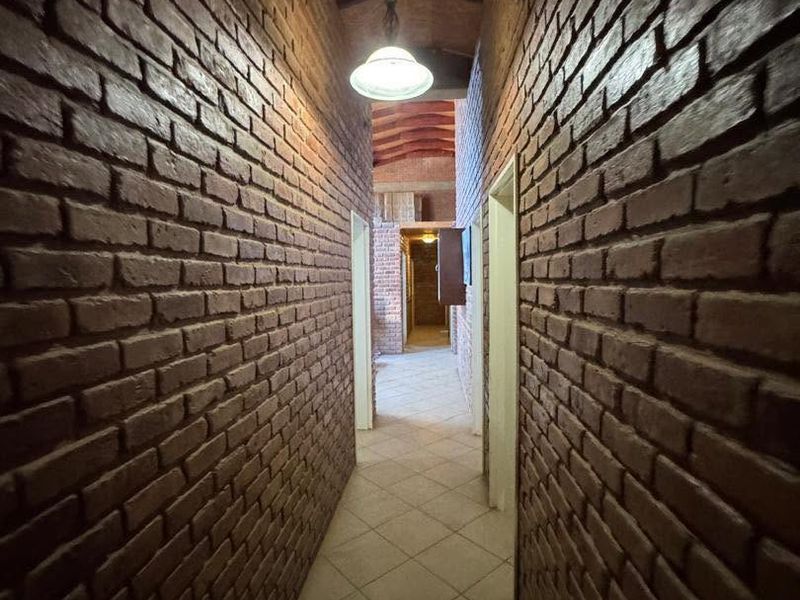 A long hallway featuring rustic exposed brick walls, a vaulted ceiling with timber beams, and neutral ceramic floor tiles.