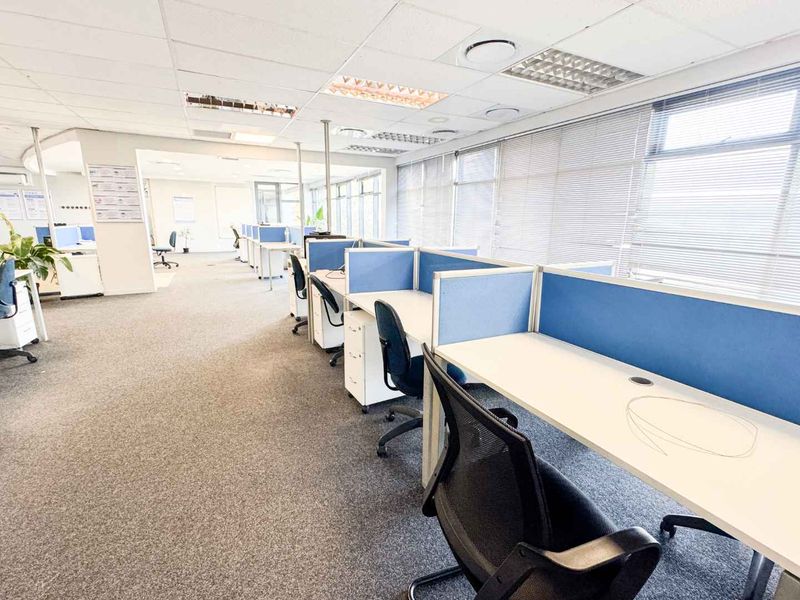 Open-plan office space featuring carpeted flooring, suspended ceilings with recessed lighting, and large windows with venetian blinds, equipped with built-in workstation partitions.