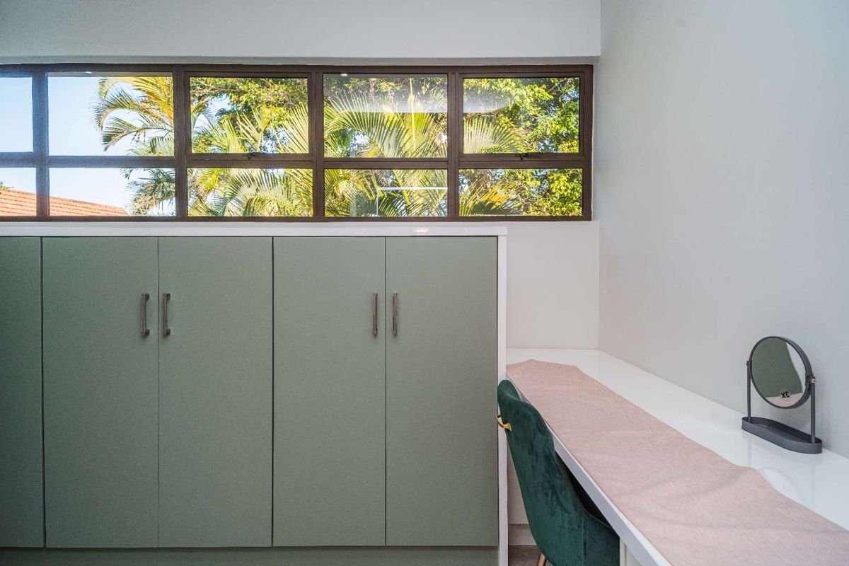 A well-lit bedroom featuring built-in sage green cabinetry and a sleek integrated desk area with large windows overlooking lush greenery.