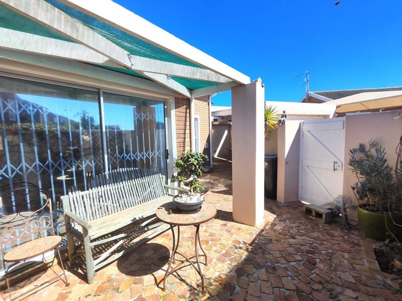 A paved outdoor patio area featuring a covered structure, sliding glass doors with security gates, and a private pedestrian access gate.