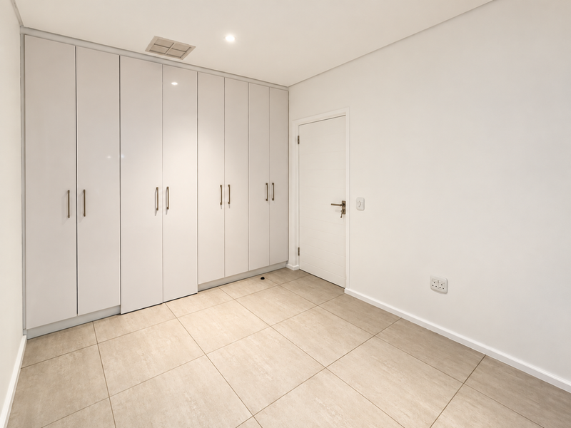 A clean, modern bedroom featuring neutral tiled flooring, ample built-in cupboard storage, and recessed lighting, presenting a bright and well-maintained space.