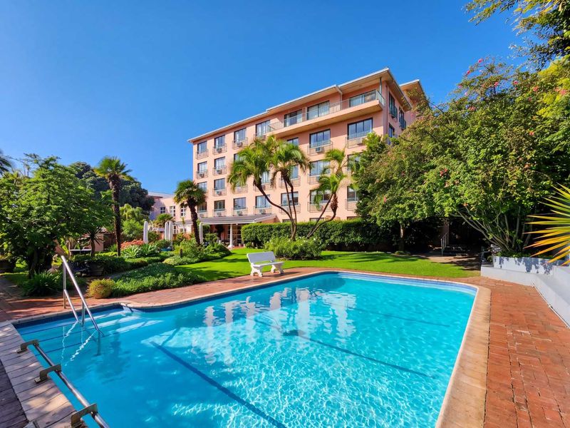 A well-maintained apartment complex featuring a clear swimming pool, brick-paved surround, and lush landscaped gardens under a bright blue sky.