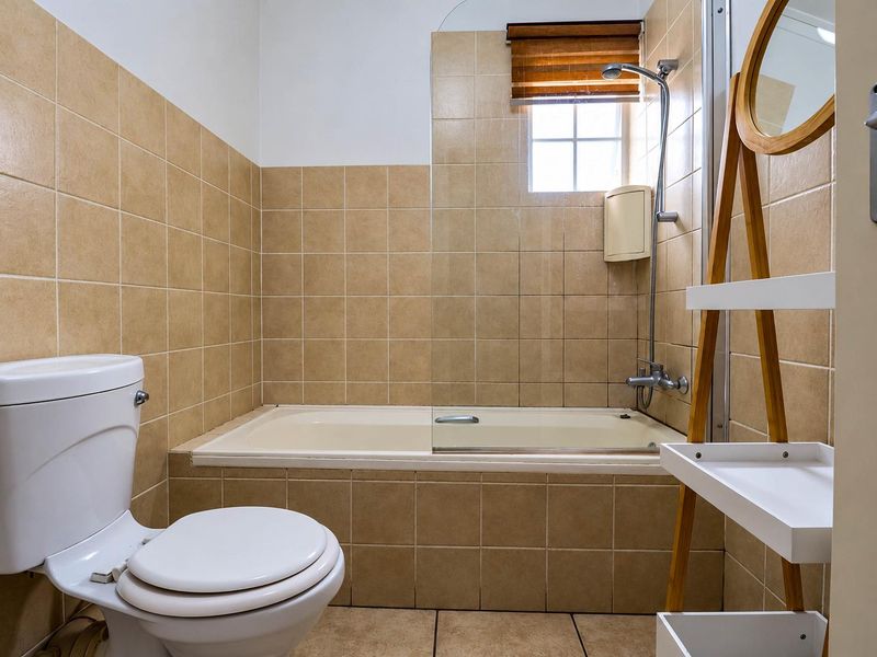 A functional bathroom featuring a built-in bathtub with a shower mixer, glass screen, and neutral ceramic tiling throughout.